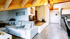 Amplia sala de estar con techo de madera alto, un sofá blanco con cojines a rayas, una mesa de comedor y una zona de cocina con armarios blancos y un microondas.