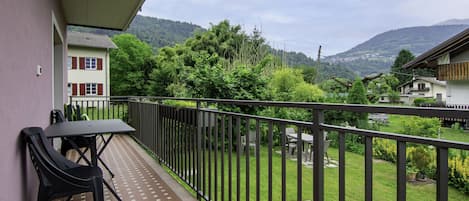 A balcony with a table and chairs, a view of a green landscape with houses, and a mountain in the distance.
