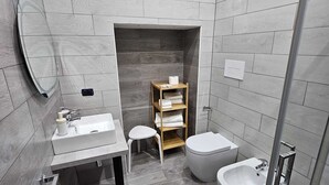 Een moderne badkamer met grijze tegels, een glazen douchecabine, een witte wastafel, een toilet en een douchetoilet.