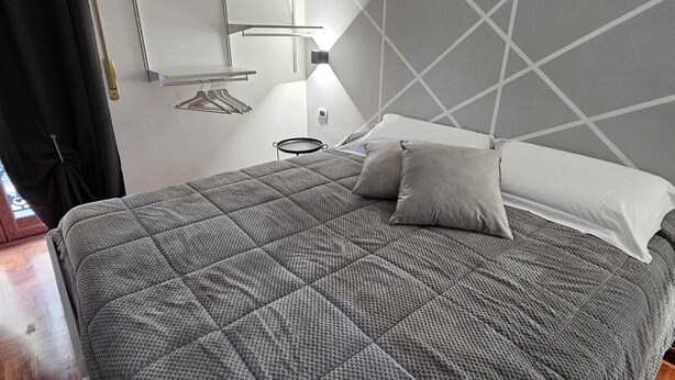 Een net opgemaakt bed met een grijze quilt, een bedlampje en een wandplank met kleding.