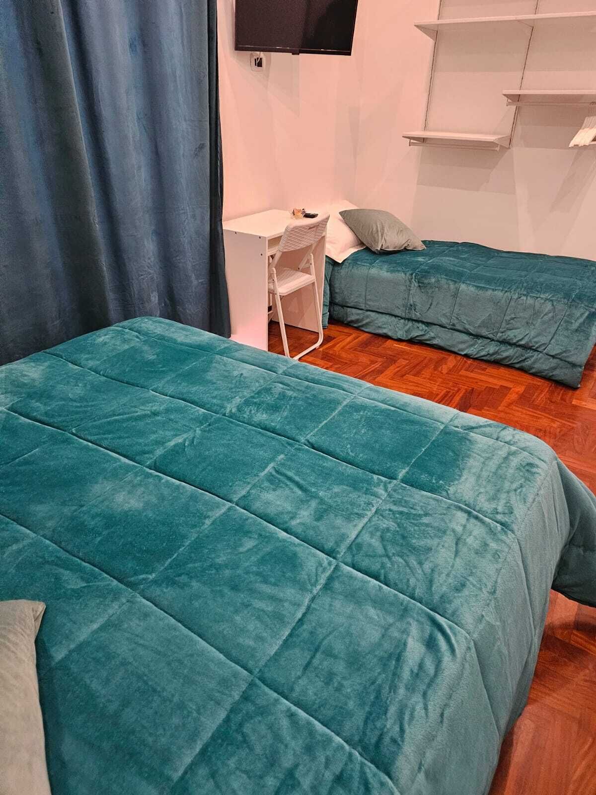 A room with two teal beds, a desk with a chair, and a TV mounted on the wall.