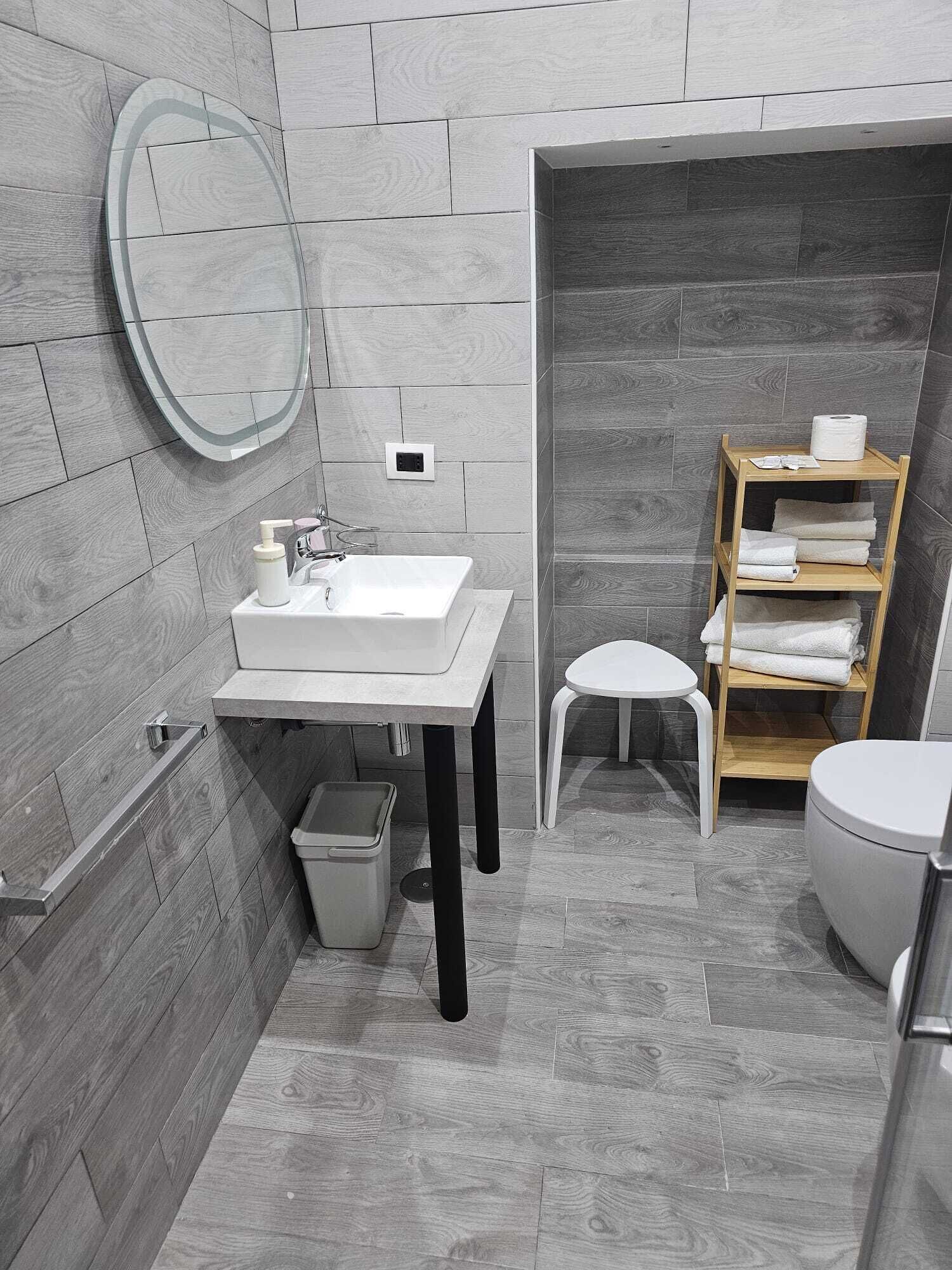 Une salle de bain moderne avec un lavabo blanc, un miroir et des toilettes. On y trouve un petit tabouret et une étagère en bois avec des serviettes.