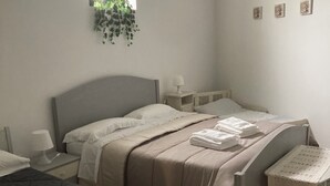 A bedroom with two beds, a bedside table with a lamp, a small window with a plant, and a wall-mounted shelf with decorative items. - The five olive trees (Alberobello)
