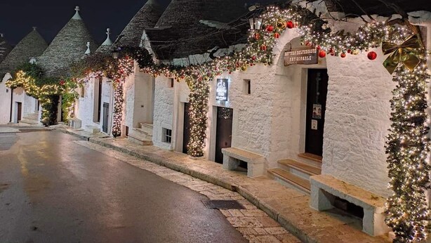A row of white buildings with conical roofs, decorated with Christmas lights and ornaments. - The five olive trees (Alberobello)