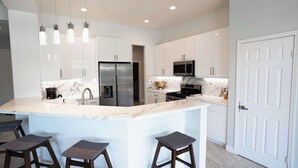 A modern kitchen with white cabinets, a marble countertop, a stainless steel refrigerator, a microwave, and a gas stove. There are three bar stools at the island. - Palm Luxe Estate: Luxury Desert Retreat Pool & Spa (Indio)