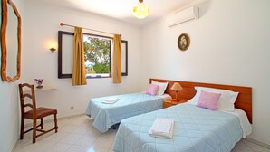 A hotel room with two beds, a chair, a window with curtains, a wall-mounted lamp, and an air conditioning unit.