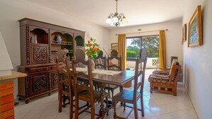 A dining area with a wooden table, chairs, and a chandelier. There is a view of the outdoors through a window.