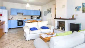 A modern living room with a white sofa, a dining table, and a kitchen area with blue cabinets and a fireplace. - Casa Bel Sit by Interhome (Acquaseria)