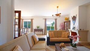A spacious living room with a sofa, a coffee table, and a fireplace. There is a dining area with a table and chairs visible through a glass door.