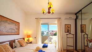 A hotel room with a bed, a nightstand, a lamp, a painting, a window with curtains, and an air conditioning unit.