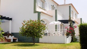 Un edificio de dos pisos con fachada blanca y detalles verdes, que cuenta con un pequeño jardín con un árbol y un paraguas blanco.