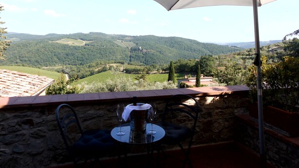 Une table avec deux chaises et un verre à vin, placée sous un grand parasol, d’où l’on peut admirer un paysage pittoresque composé de collines ondoyantes et d’un village situé au loin.