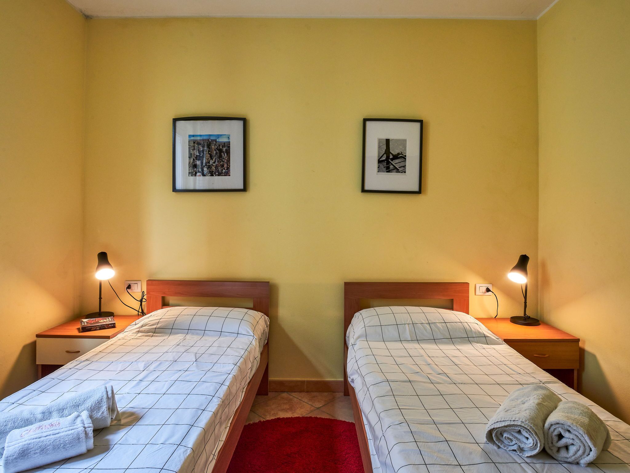 A hotel room with two beds, each with a wooden headboard, a red rug, and two bedside lamps. There are framed pictures on the wall above the beds.