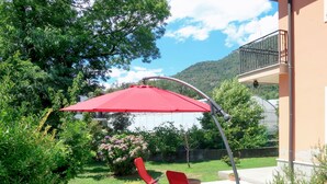 A patio with two red chairs and a red umbrella, surrounded by lush greenery and a view of a mountain. - casa dei fiori by Interhome (Mergozzo (Lago di Mergozzo))