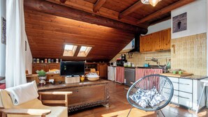 Un espacio habitable acogedor en el ático, con techo de madera, televisor, sofá, mesa de comedor y zona de cocina con armarios de madera y salpicadero de azulejos.