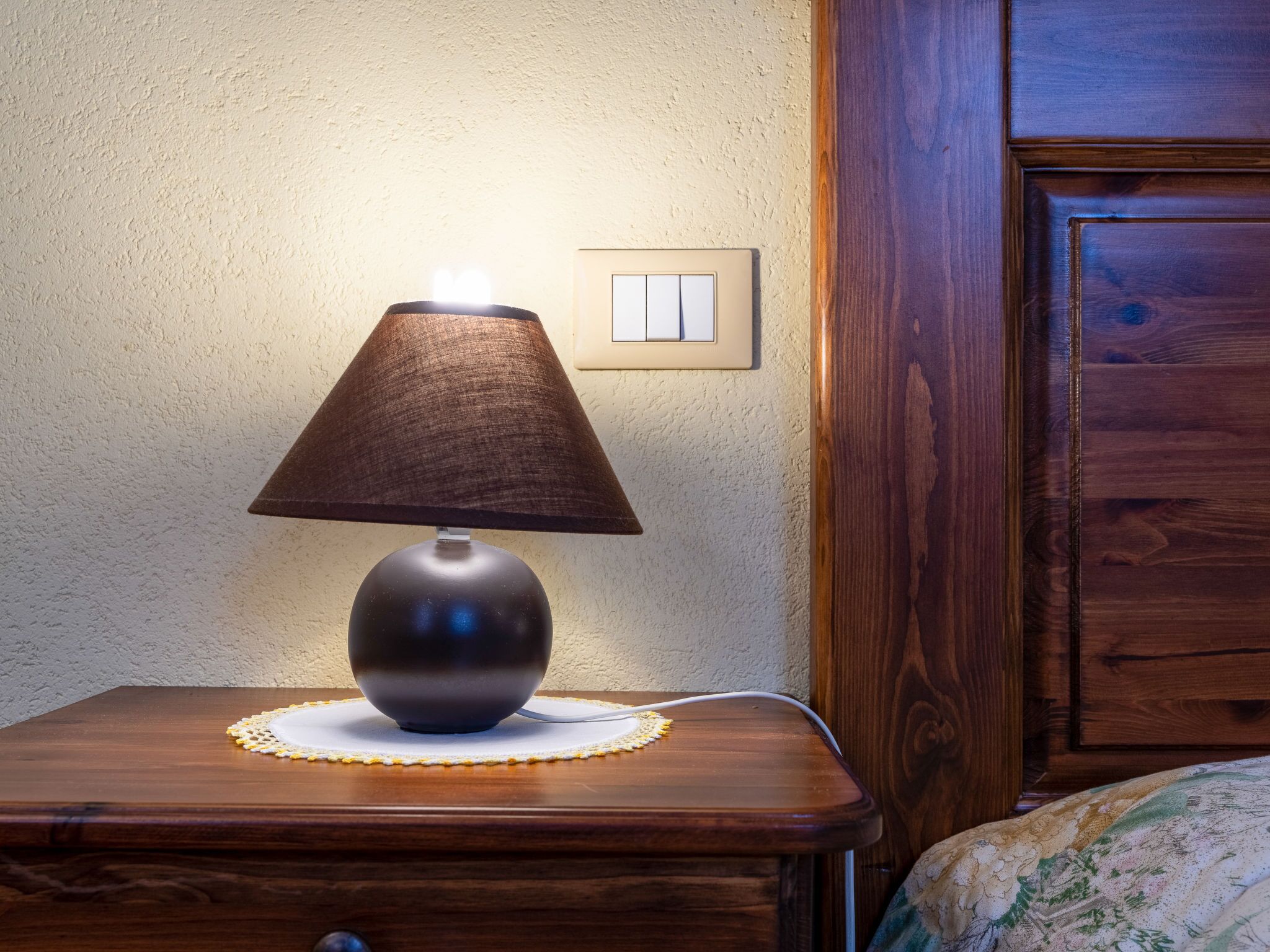 A bedside table with a lamp, a power outlet, and a wooden cabinet in the background.