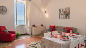 Un coin repas avec une table dressée pour quatre personnes, un fauteuil rouge, un canapé avec des coussins rouges, une horloge au mur et une vue sur la mer par la fenêtre.