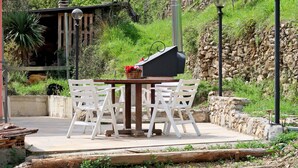 An outdoor patio with a wooden table and white chairs, surrounded by a stone wall and greenery. - Oasi Colorata by Interhome (Stellanello)