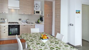 A modern kitchen with wooden cabinets, a microwave, and a sink. There is a dining table with a floral tablecloth and a bowl of fruit. - La Perla del Mediterraneo by Interhome (Cervo)