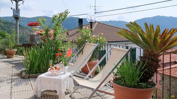 Um terraço com uma mesa posta para dois, rodeado por plantas em vasos e uma cadeira.
