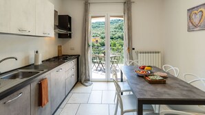 A modern kitchen with a gas stove, a sink, and cabinets. There is a dining table with chairs and a view of a green landscape through a glass door. - Casa Mimosa by Interhome (Pontedassio)