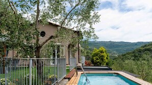 A swimming pool with a wooden deck, surrounded by a metal fence, and a building with a red roof in the background. - Casa Mimosa by Interhome (Pontedassio)