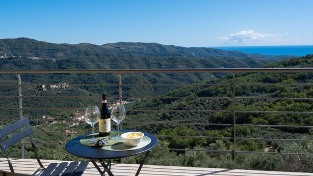 A table with a bottle of wine, glasses, and a bowl of food on a terrace overlooking a mountainous landscape. - Bellaria (VLO190) by Interhome (Valloria)