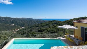 A pool with a clear view of the surrounding landscape, a dining area with yellow chairs, and a building with a tiled roof. - Bellaria (VLO190) by Interhome (Valloria)