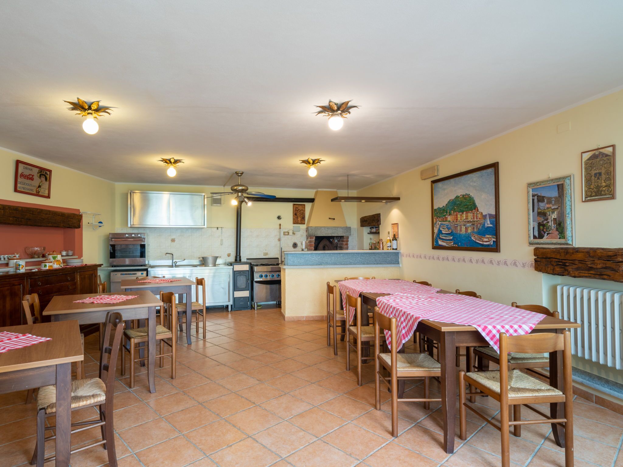 A dining area with wooden tables and chairs, a kitchen in the background, and a painting on the wall.