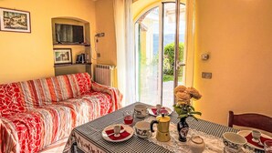 A dining area with a checkered tablecloth, a sofa with a red and white patterned cover, and a view through a glass door to an outdoor area with greenery. - Primula (VLO206) by Interhome (Valloria)