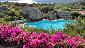 Ein Pool, umgeben von großen Felsen und rosa Blumen, mit Blick auf das Meer und ferne Berge.