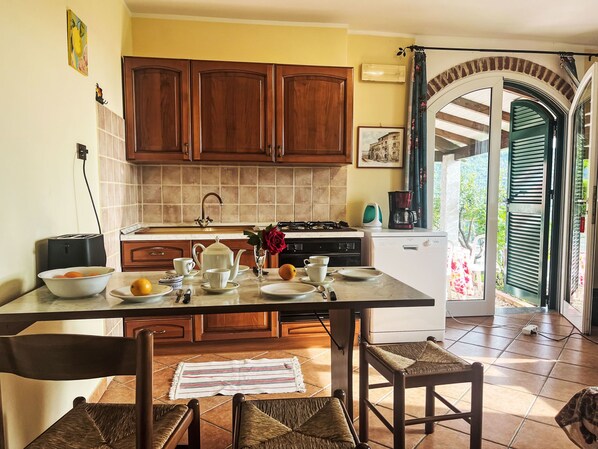 Una cocina con armarios de madera, salpicadero de azulejos, un fregadero, una estufa y una cafetera. Hay una mesa de comedor con sillas y un jarrón con flores.