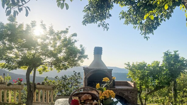 Un cadre extérieur rustique avec une cheminée en pierre, une table en bois agrémentée d’un panier de pain et de fleurs, et une vue sur des montagnes lointaines.