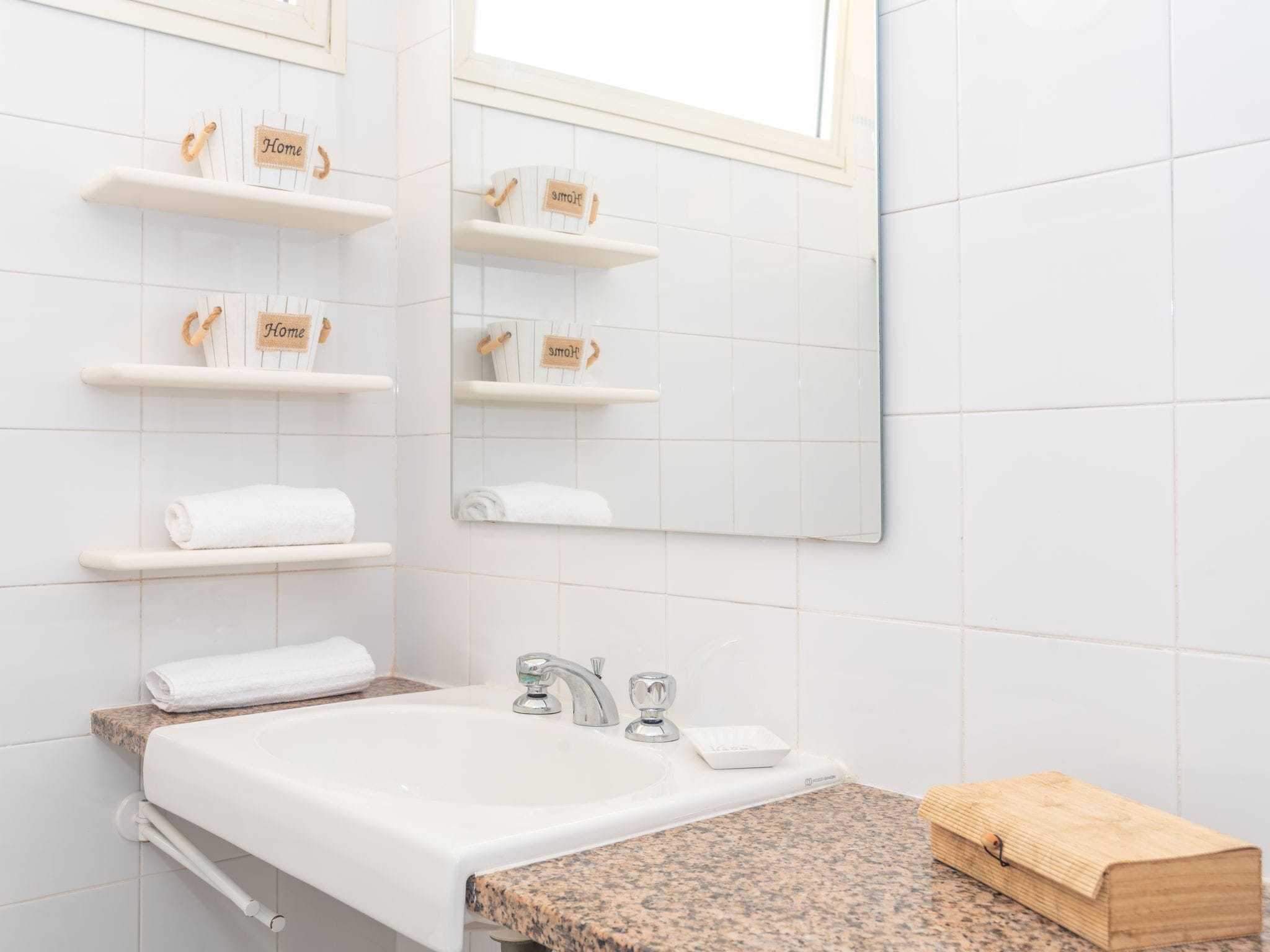 Une salle de bain avec des carreaux blancs, un lavabo avec deux robinets et des étagères où sont posées des serviettes, ainsi qu’un panneau indiquant « Chez soi ».