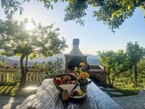 Entorn rural a l'aire, amb una llar de pedra, taula de fusta, cistella de pa i flors, i vista a muntanyes llunyanes.