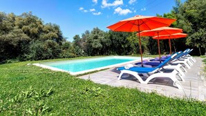 A swimming pool with lounge chairs and red umbrellas, surrounded by greenery. - Oliviola by Interhome (Valledoria)