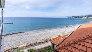 Blick auf die Küste mit einem Kiesstrand, einem Gebäude mit rotem Dach und einer weiter entfernten Stadtansicht.