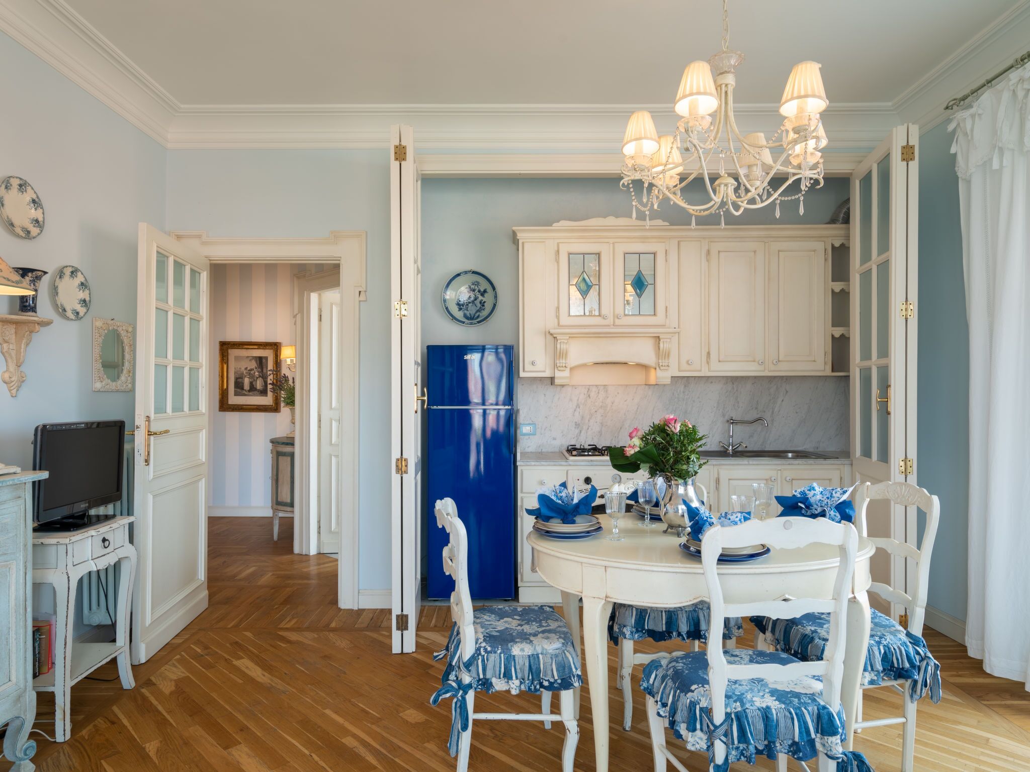Un comedor con una mesa y sillas blancas, una araña y un refrigerador azul.