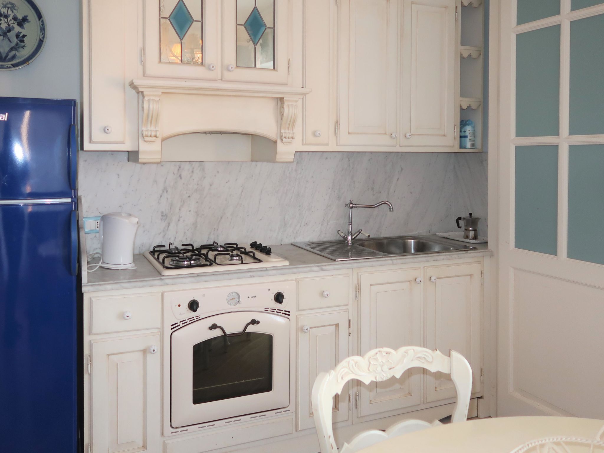A kitchen with white cabinets, a blue refrigerator, a gas stove, a sink, and a white chair.