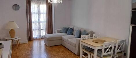 A modern living room with a sofa, a dining table with chairs, a lamp, and a large window with curtains.