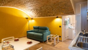 Un espacio habitable compacto con techo de ladrillo, sofá color esmeralda, una pequeña mesa de comedor y una kitchenette.