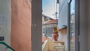 Un pequeño balcón con una silla, una mesa con un florero y copas de vino, y vistas a un edificio con tejas rojas.