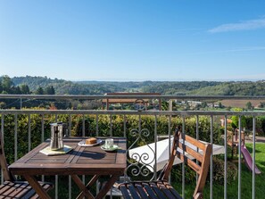 A wooden table with a metal chair, a metal pitcher, a plate with a slice of cake, and a cup on a balcony overlooking a scenic landscape with trees and buildings. - Grappolo + Margherita by Interhome (Cossombrato)