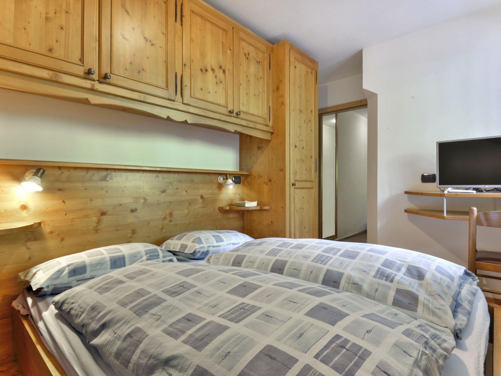 Une chambre à coucher avec une tête de lit en bois, un lit recouvert d’une couette à carreaux, une lampe de chevet et un téléviseur posé sur un meuble en bois.