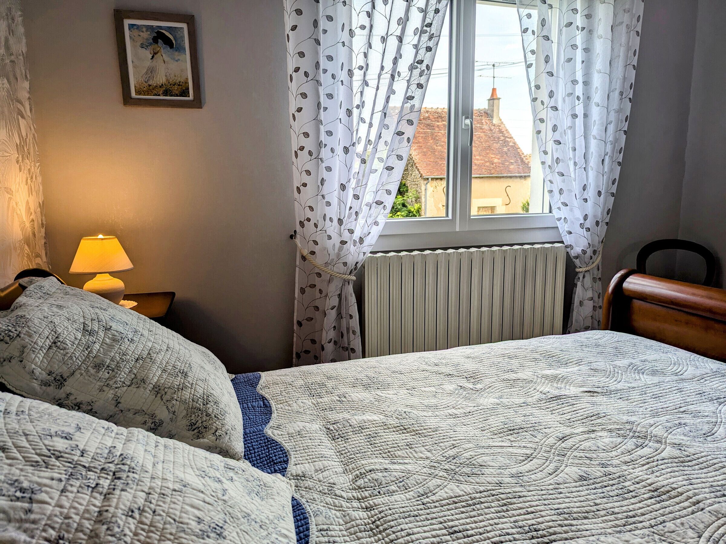Een slaapkamer met een bed, nachtkastje met lamp, raam met gordijnen, radiator en een schilderij aan de muur.