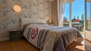 A bedroom with a large bed, a nightstand, a wall-mounted light, and a view of the outdoors through a window with curtains. - Renovated Townhouse in Argy: Private Garden, Game Room, Near Château and ZooParc de Beauval (Argy)
