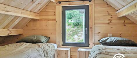 A wooden cabin with two beds, a small table, and a window with a view of greenery.
