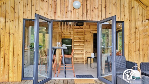 Une cabane en bois avec une porte d’entrée en verre, un coin repas comprenant une table et des chaises, et un canapé en osier.