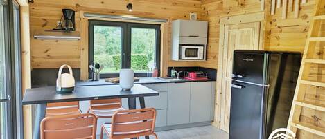 Une cuisine compacte aux murs en bois, avec un réfrigérateur noir, un four à micro-ondes et un évier. On y trouve deux chaises orange et une table avec une bouilloire.