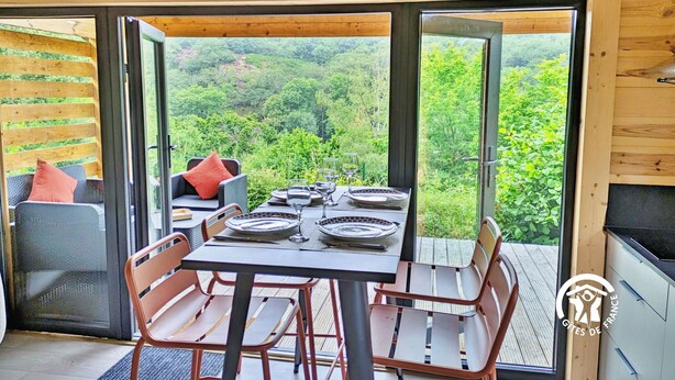 Comedor tipo loft con una mesa dispuesta para cuatro personas, rodeada de sillas, y vistas a un bosque frondoso a través de amplias puertas de cristal.